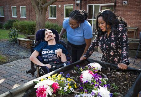 Person supported in wheelchair with two colleagues in garden.