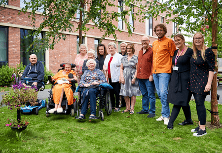 A group of people supported at the Watch Factory in Prescot, smiling for a photo in the garden with Photographers in residence, Sam and Marge.