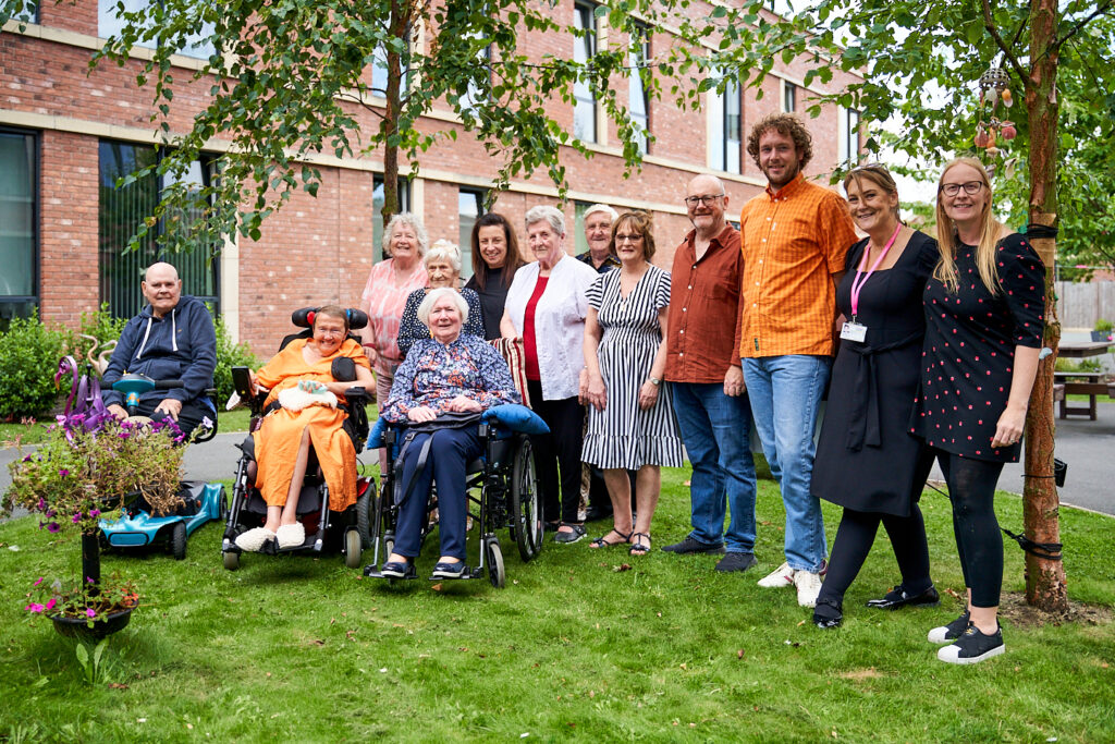 A group of people supported at the Watch Factory in Prescot, smiling for a photo in the garden with Photographers in residence, Sam and Marge.