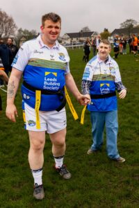 Two Learning Disability Super League players standing on the pitch looking at the camera. One wearing a blue and white rugby top and white shorts, one man wearing a blue and white jacket.