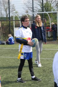 Jake holds a rugby ball, wearing a white rugby top and blue shorts with black leggings underneath. He stands on a rugby pitch, looking to his teammates.