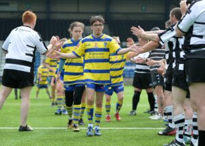 A line of players wearing blue and yellow striped rugby tops, led by Jake, walk through two lines of players in black and white stripes rugby tops. The players from opposite teams are high five-ing each other as they pass.
