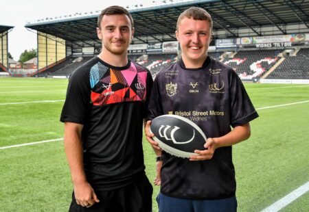 Two men wearing black Widnes Vikings shirts pose for the camera in front of the Vikings Stadium, with the pitch behind them.