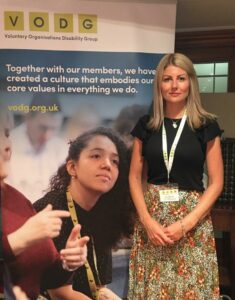 Director Jen Moores stands in front of a VODG advertisement banner. She is wearing a lanyard and is smiling to the camera.