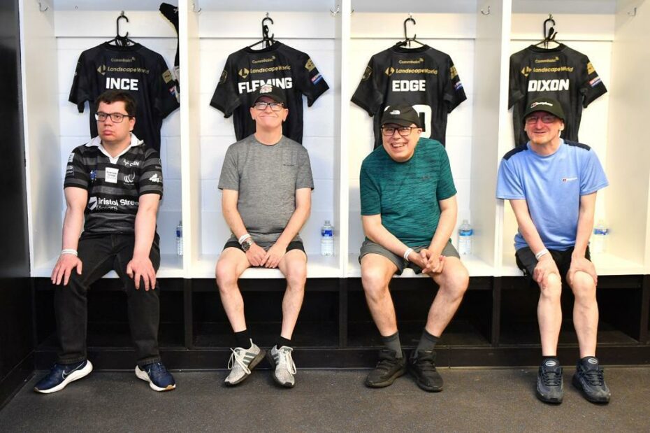 Four people supported by Community Integrated Care sit in front of Widnes Vikings' player's t-shirts, hung up in the stadium changing rooms, as part of their VIP experience.