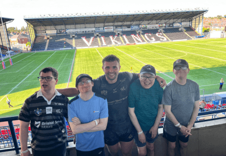 Four people supported and a colleague from Community Integrated Care stood smiling, up in their viewing box at Widnes Vikings stadium with the pitch below behind them. Photo taken by Ste Jones.