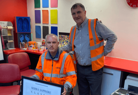 Barry and his colleague, Neil smiling to camera. Both are wearing orange high vis jackets with the Algeos logo on the front.