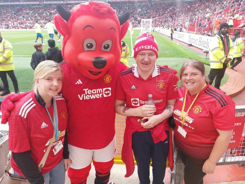 Nigel wearing a Manchester United shirt, with two support workers also wearing fan shirts and posing with the team mascot.