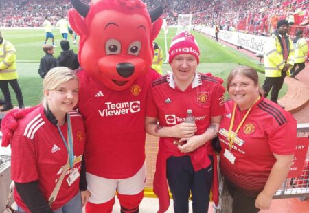 Nigel wearing a Manchester United shirt, with two support workers also wearing fan shirts and posing with the team mascot.