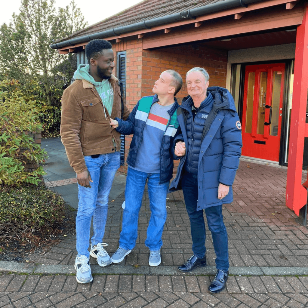 Support Worker (Omolulu), person supported (Philip) and Wallace Dobbin, Community Integrated Care Trustee) stand outside in front of Donald Dewar Court, smiling to camera. The visit took place as part of Trustee Week celebrations.