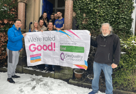 Colleagues and people supported at Norfolk Road Care Home stand outside the building with ig smiles. They are holding a banner which reads 'We're rated Good!'