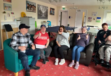 People we support sit on sofa and comfy chair, giving a thumbs up to the camera