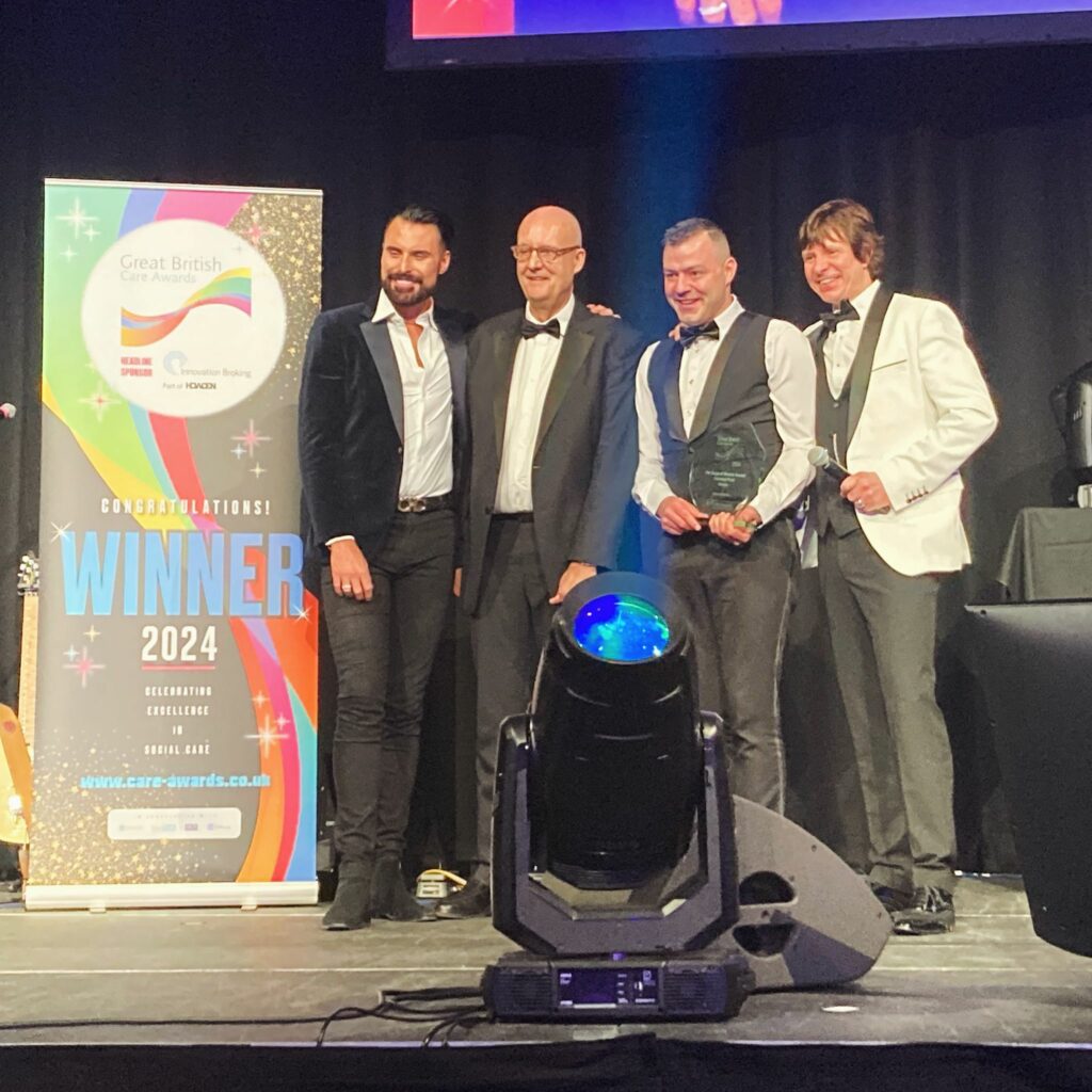 Phill stands onstage with Awards hosts and special celebrity guest, Rylan Clark. All are wearing suits and stood next to a Winners banner. Phill smiles as he holds his award for Support Worker of the Year.