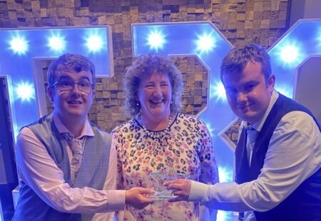 Elaine stands smiling with the people she supports, Kevin and Robert, at the Moray & Banffishire Heroes Awards, all holding the trophy for Carer of the Year.