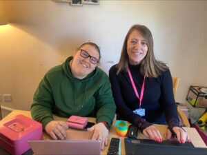 Chloe (left) and Recruitment Specialist, Natasha Palliser (right), sit together at a desk. They have their laptops on the table and they are smiling to the camera.