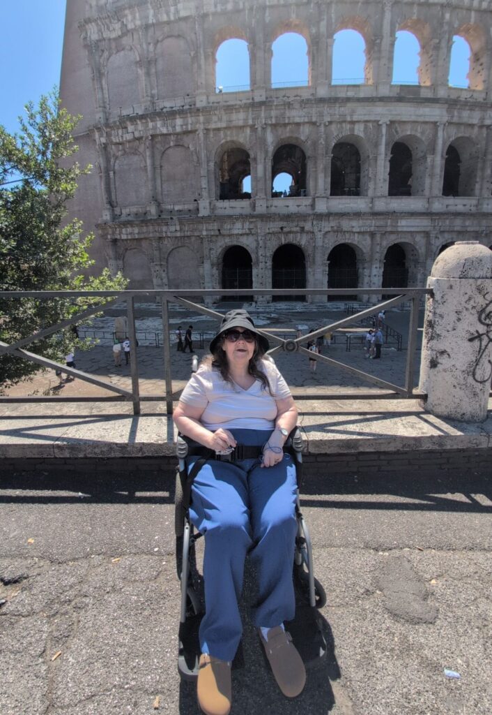 Lisa smiles in front of the colosseum in Rome. She is sat in her wheelchair wearing a navy bucket hat, white t-shirt, blue jeans and sunglasses.