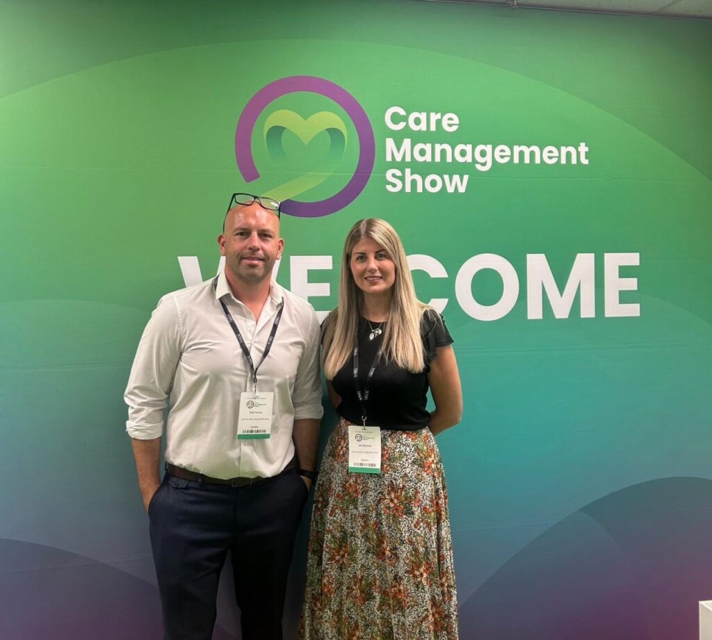 Edd and jen stand smiling to camera with event lanyards on. They're stood in front of a green, blue and purple background with 'Care Management Show' and a house logo on it.