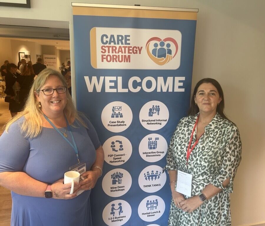 Joanna Scammell and Samantha Brennan stand smiling on either side of a ‘Welcome’ event banner for the Care Strategy Forum. The banner features the forum’s logo at the top, with icons and text highlighting the different themes of the event.