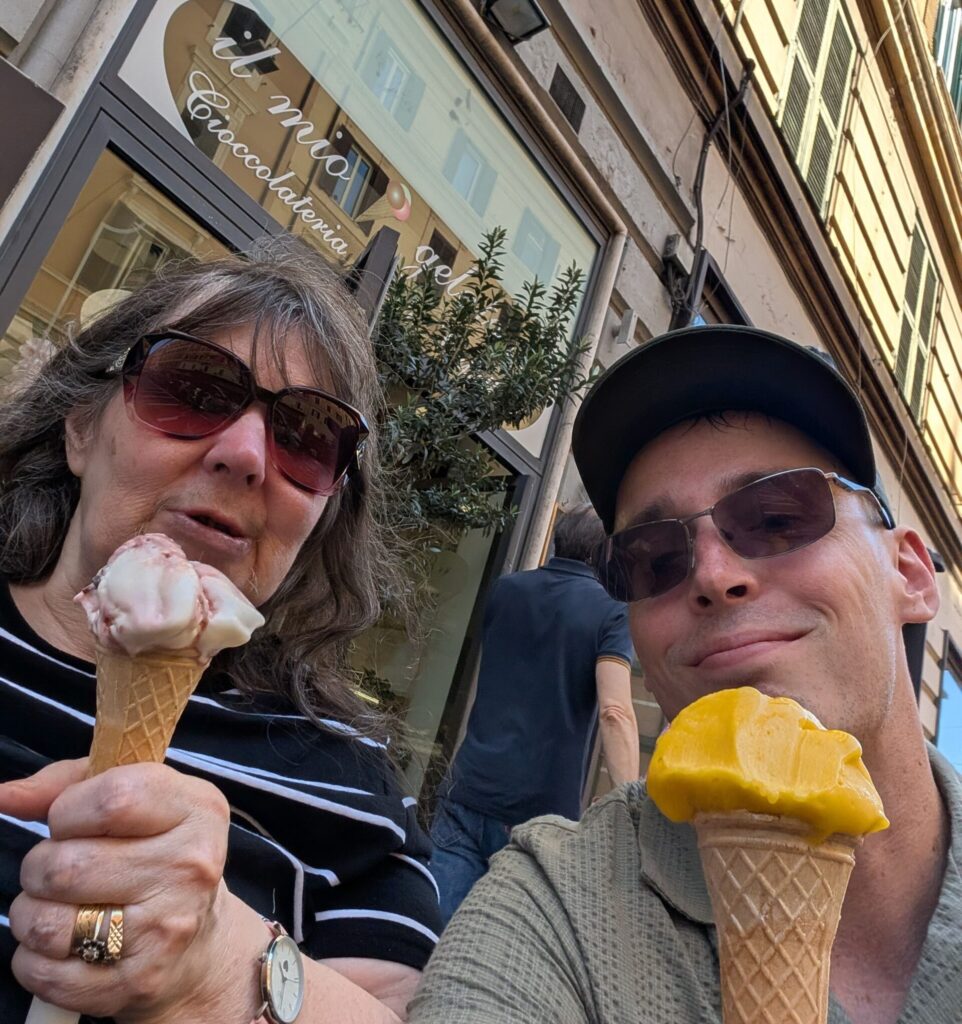 Lisa on the left smiles to camera with a pink ice cream and Scott is on the right, smiling to camera with a yellow ice crea. Both have sunglasses on and are outside a shop front in Rome.