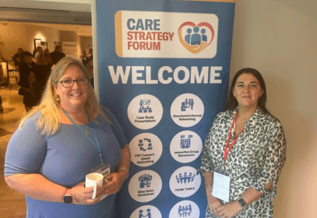 Joanna Scammell and Samantha Brennan stand smiling on either side of a ‘Welcome’ event banner for the Care Strategy Forum. The banner features the forum’s logo at the top, with icons and text highlighting the different themes of the event.