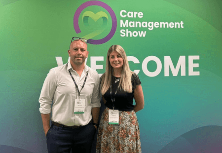 Edd and Jen stand smiling to camera with event lanyards on. They're stood in front of a green, blue and purple background with 'Care Management Show' and a house logo on it.