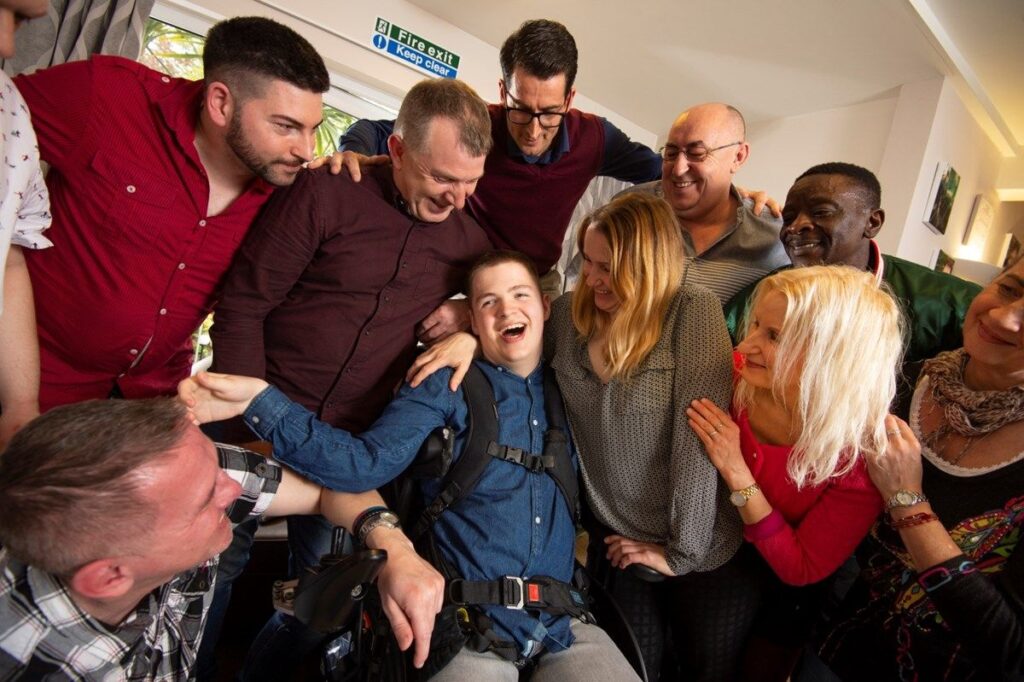 Colleagues huddle around a person supported in a wheelchair. Everyone is laughing and smiling at one another.