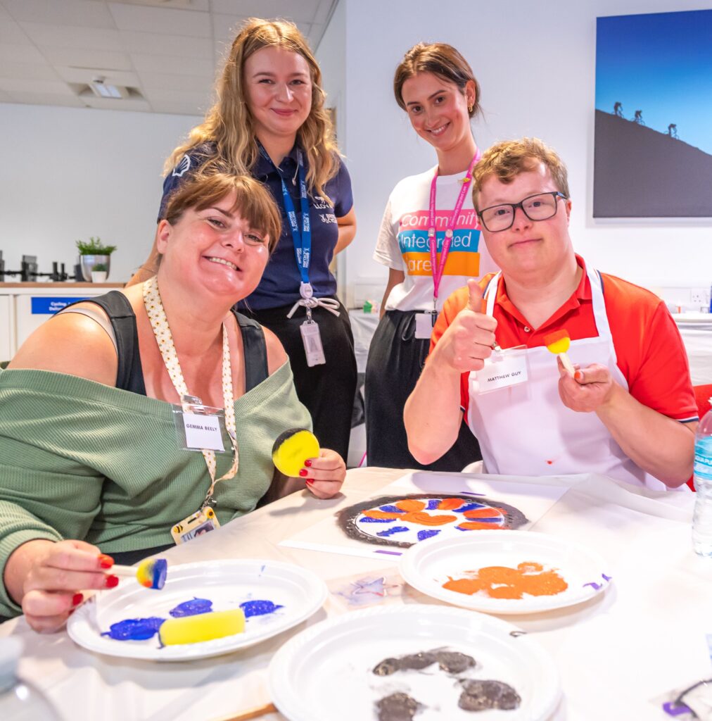 Colleagues from Community Integrated care, a person supported and a colleague from British Cycling all at a table, smiling to camera. In front of them is paint, stencils and the artwork that they are working on.