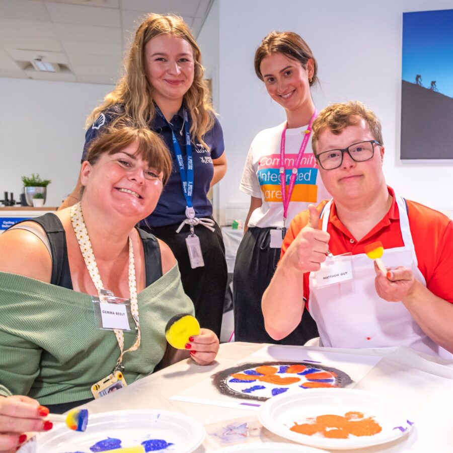 Colleagues from Community Integrated care, a person supported and an athlete from British Cycling smile to camera. In front of them is paint, stencils and the artwork that they are workign on.