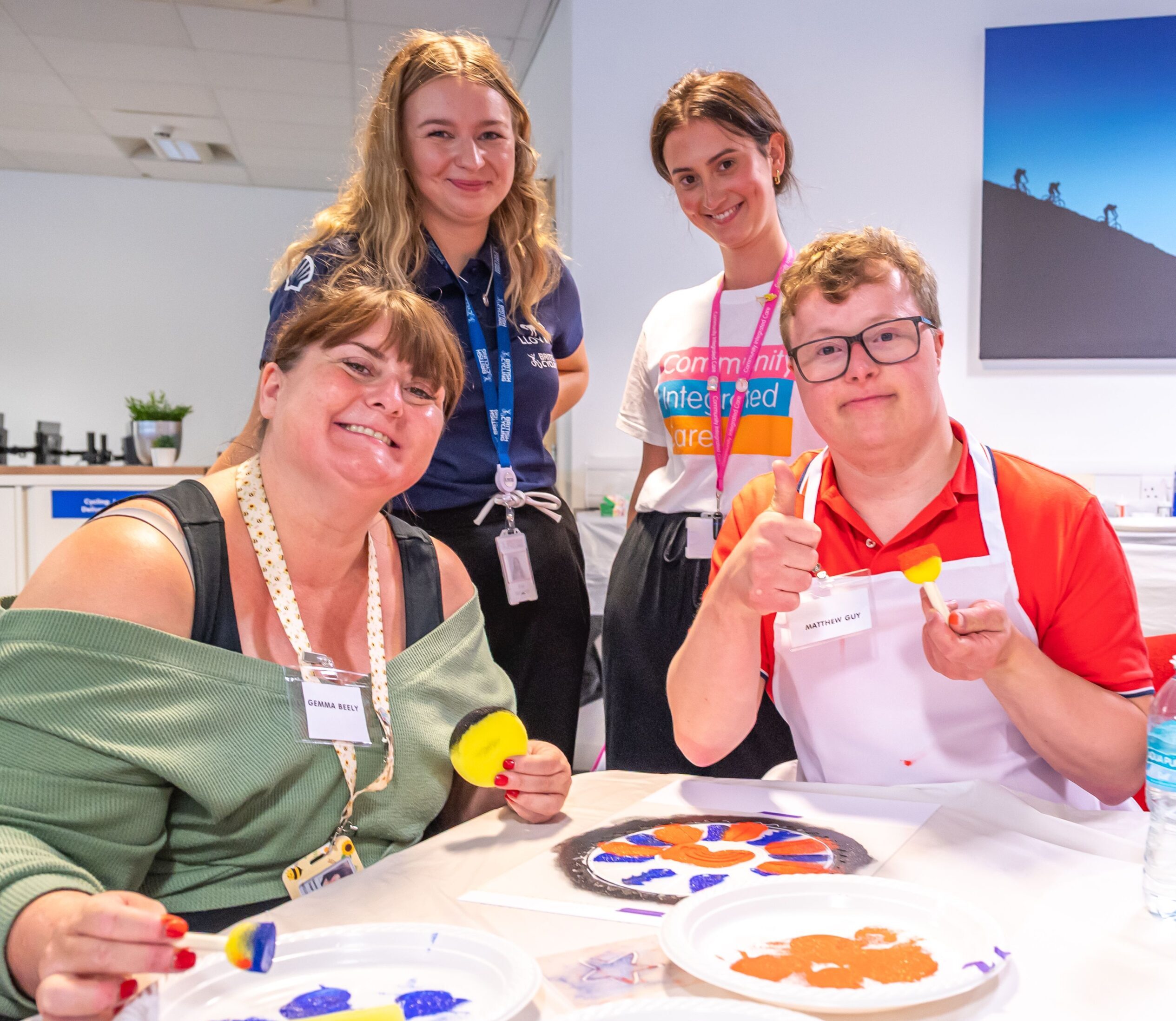 Colleagues from Community Integrated care, a person supported and an athlete from British Cycling smile to camera. In front of them is paint, stencils and the artwork that they are workign on.
