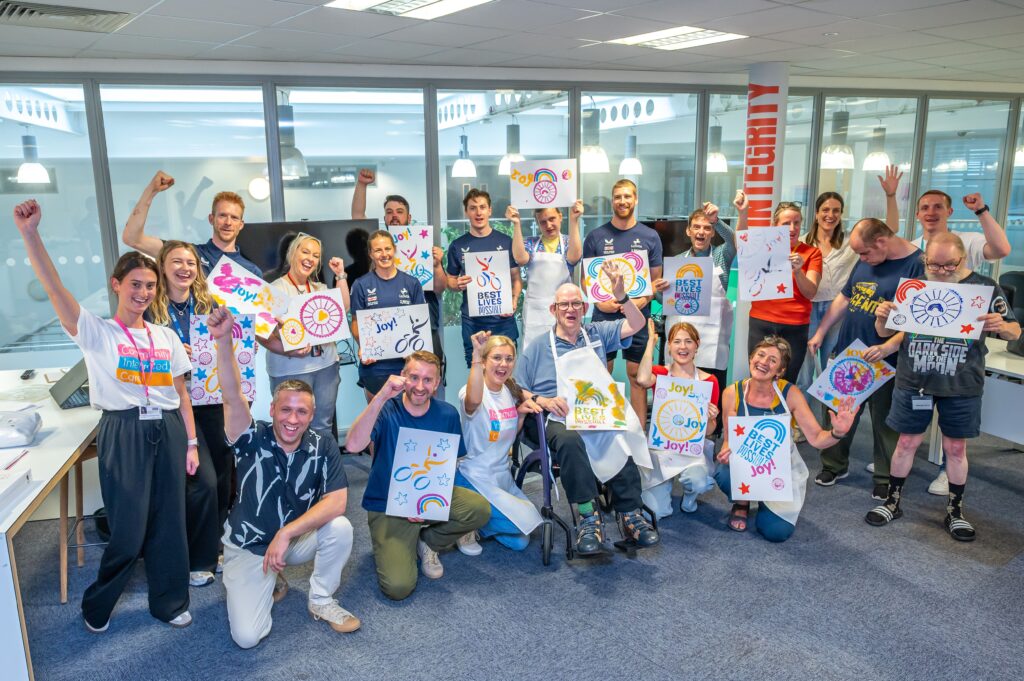 A group of colleagues from British Cycling and colleagues and people supported by Community Integrated Care smile and cheer to camera, holding stencil paintings. The paintings are in an assortment of colours and feature different icons, including cyclists and rainbows. 