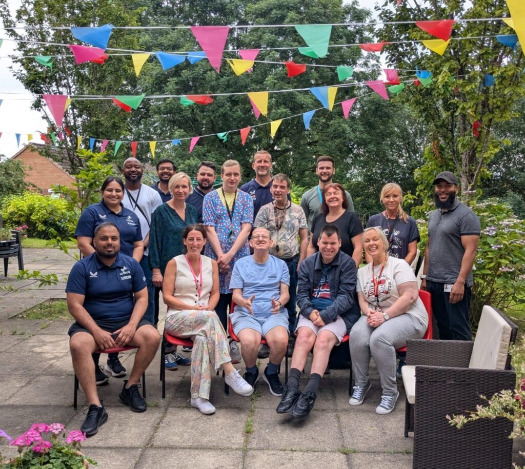 A large group of colleagues and people supported with officials from British cycling stand for a group picture. They're outside on the patio of a garden surrounded by trees strung with rainbow bunting.