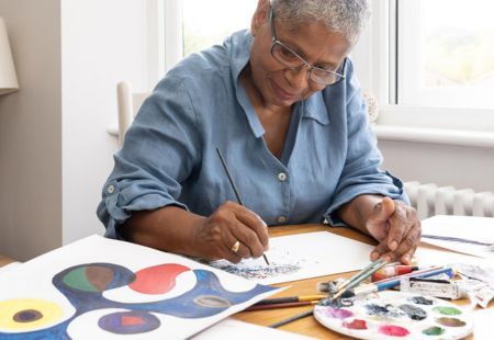 A person sits at a table with a paintbrust and palette. They're painting with vibrant colours.