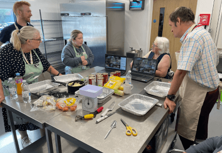 Ed Clancy stands in a kitchen with four people supported by Community Integrated Care. They are surrounded by cooking ingredients and utensils. They have a laptop in front of them and they are watching a live cook along, hosted by Rosemary Shrager.