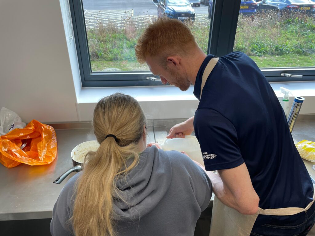 The photo is taken from behind Ed Clancy and a person supported by our charity. Ed is helping pour ingredients into a mixing bowl.