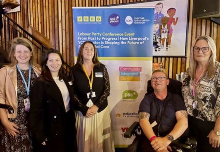 Chris, a person supported by Community Integrated Care, smiles to camera. Stood next to hims is Marie Godfrey, Regional Manager, Sam Brennan, Managing Director, Danielle Chan, Director of Communications and Imogen Shillito, Trustee. Behind them is a pull up banner about the event, with a Community Integrated Care logo included.