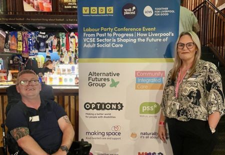 Chris, a person we support, and Marie, a Regional Manager stand in front of an events banner, smiling to camera.