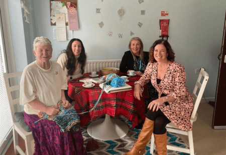 A person we support (Susan) sits at her dining table with her knitting. She is joined by our Head of Operations, Caroline, and two Trustees, Sue and Kathryn.