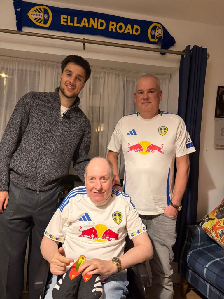 Ilia stans next to Leslie and his support worker, Stephen, all smiling to camera. Hanging up on the wall above them is an Elland Road scarf.
