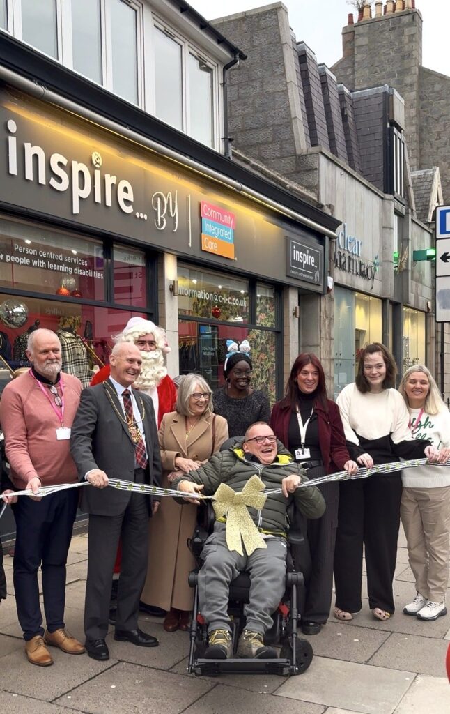 Colleagues and people supported by Community Integrated Care stand outside the Inspire By shop with the Lord provost of Aberdeen. They are all holding sparkly ribbon. Maurice (centre) is getting ready to cut the ribbon.