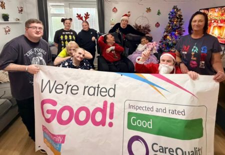 Colleagues and people supported at Bentinck Crescent stand together, holding up a banner that reads 'We've been rated Good by CQC'. The room they are in is decorated for Christmas with sparkling streamers and a Christmas tree and they are all wearing Christmas attire, including Santa hats and reindeer antlers.