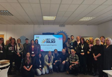 A group of senior leaders from Community Integrated Care stand smiling to camera, around a screen which reads 'PBS Senior Leaders Programme'.
