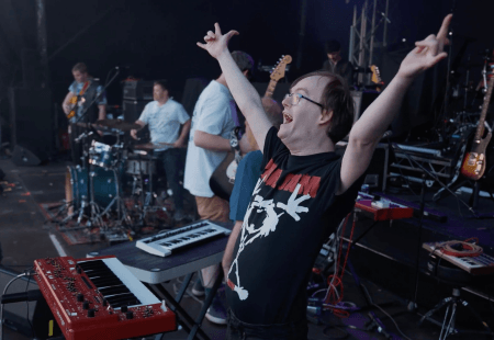 A musician from Ultimate Thunder stands at the front of a stage with arms raised triumphantly in a rock‑and‑roll pose. A keyboard and other electronic equipment sit on a table in front of them. Behind them, band members play drums, guitars and other instruments as on a dimly lit stage filled with musical gear and cables.