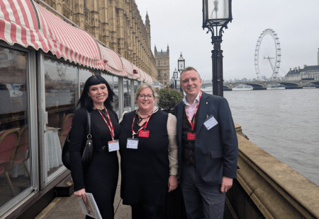 Three people wearing conference badges stand by the River Thames with the London Eye and Westminster Palace in the background.