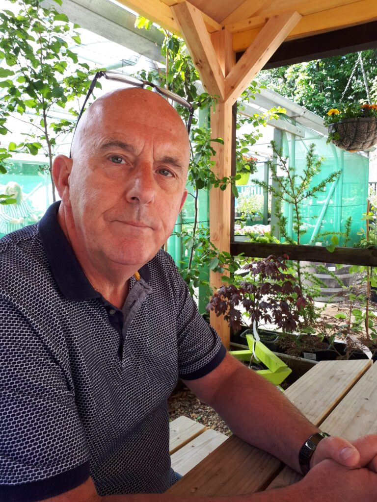 John Scott, sat on a picnic bench outside, smiling to camera. There are plants behind him.