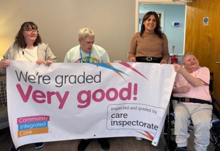 Colleagues and people supported at Muirs Court are smiling and holding up a banner reading 'We're rated Very Good by the Care Inspectorate'.