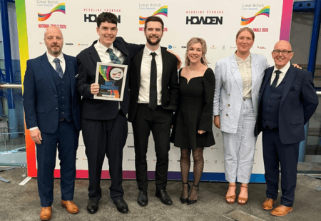Six people stand in front of a GReat British Care Awards backdrop, smiling to camera. Terry, a person supported by the charity, stands in the middle holding a Highly Commended certificate for What To Do.