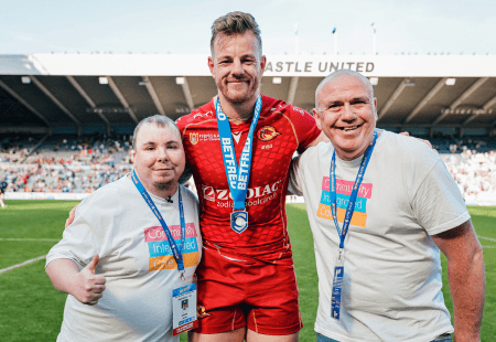 A Rugby League player in a red sports kit stands on a rugby pitch with two people supported by our charity, wearing white Community Integrated Care T-shirts, on either side. All three have event lanyards around their necks, and one of the people in white is giving a thumbs‑up. The stadium seating and spectators are visible in the background.