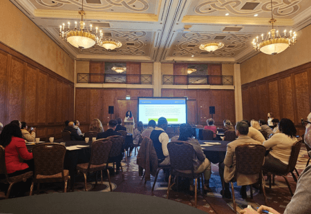 “A conference room at the Complex Care Forum with ornate chandeliers and wood‑panelled walls, where attendees sit at round tables facing a presenter at the front. A large screen displays a presentation slide, and the audience is listening while seated throughout the room.