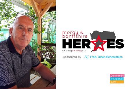 John Scott, sat on a picnic bench outside, smiling to camera. There are plants behind him. Next to John's photo is a logo that says 'Moray and Banffshire Heroes 2026'