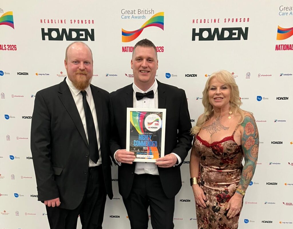 Three colleagues from Griffin Lodge are stood in front of a Great British Care awards backdrop. Service Leader, James, is stood in the middle, holding a Highly Commended certificate. They are all smiling to camera.